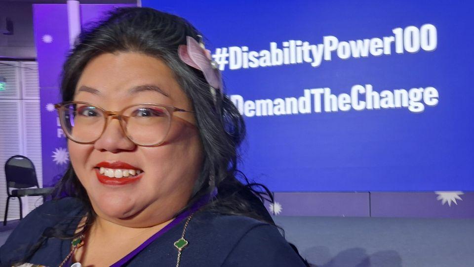 A woman is photographed infront of a screen which reads 'disability power 100'