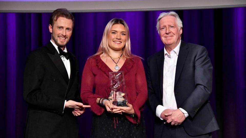 Graduate Sophie Sutcliffe holding the glass trophy in the centre with two people presenting the award.