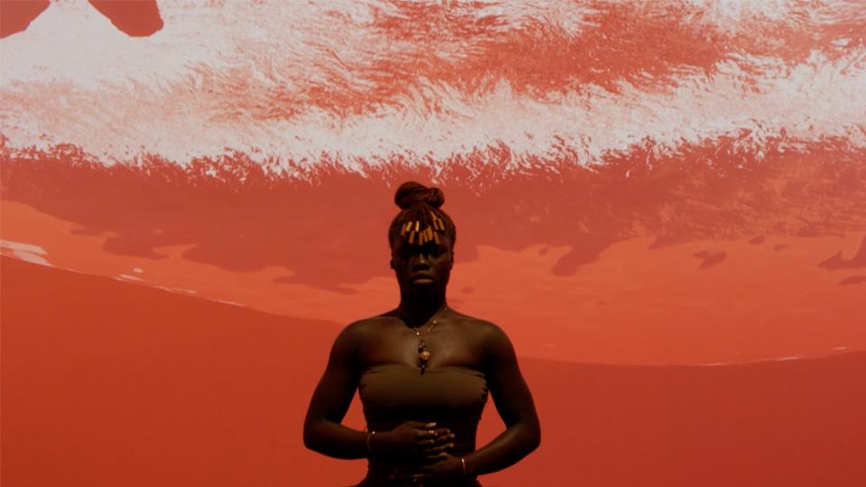 A performer stands with their hands on their diaphragm, against a red and white abstract projected image.