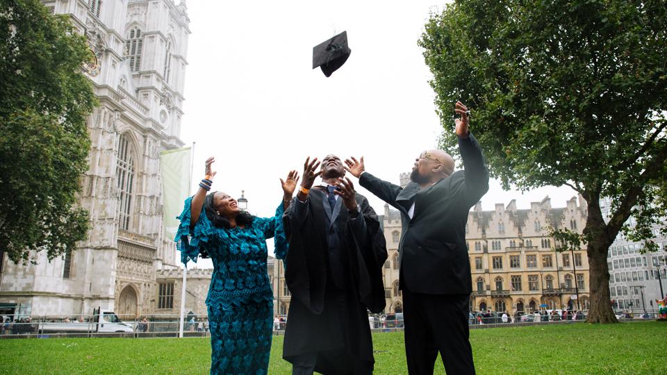 Matthew, a Black man, throws his graduation cap in the air in front of Westminster Abbey. He is standing between his parents.