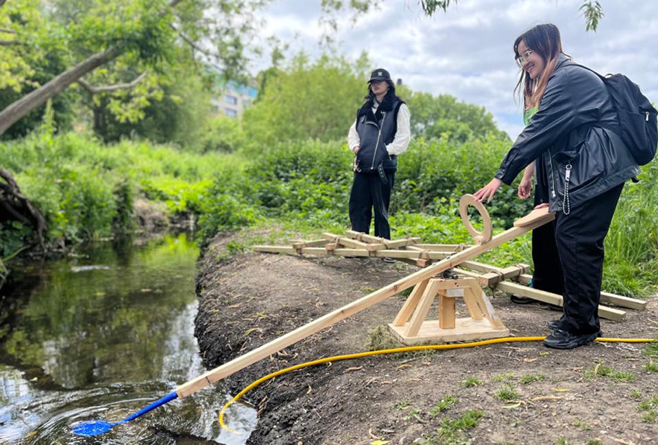 Two young women stand by a river, one holds a large wooden rod in the water