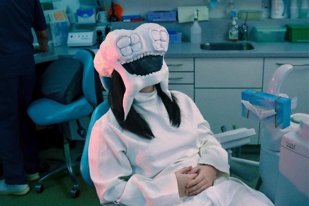 A woman wearing a mask of a clean white tooth site in a dentists chair. The environment is clear and clinical