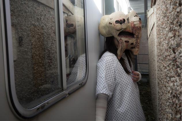A woman wearing a mask of a mostly decayed tooth stands in a narrow passageway between a caravan and a wall