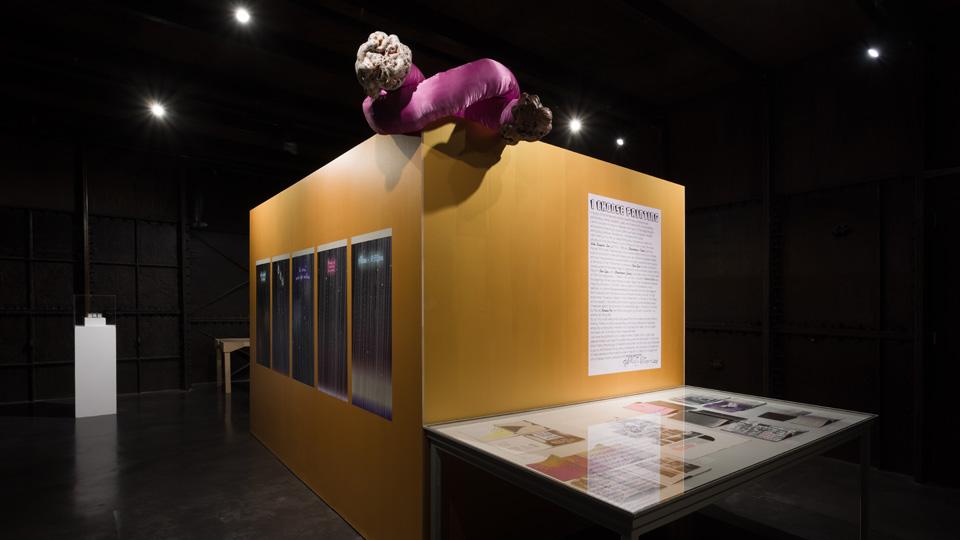 A gold plinth in a dark gallery space with a cartoon-like purple sculpture on to