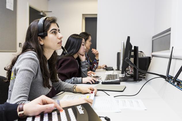 Three students sit at the gallery that forms part of the TV studio – the student in the foreground wears a headphone and mic and holds a script in their hand