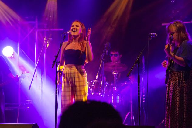 A music student stands in front of a microphone, singing. There are two bandmates in the background – one on drums, and one playing a guitar.